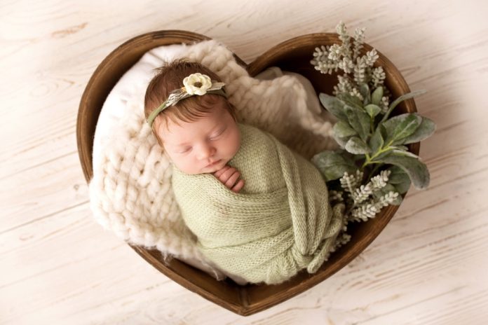 A cute newborn baby in a green pistachio winding and a white bandage with a flower on her head sleeps sweetly in a Wooden basket in the shape of heart.