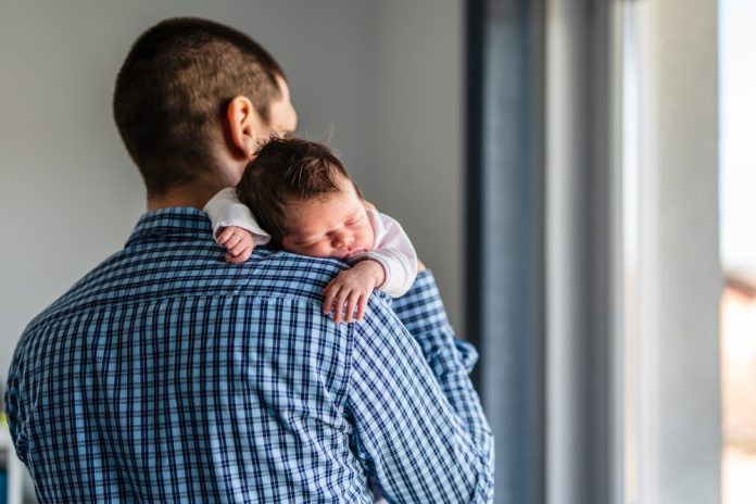 Back view of father holding his newborn baby while standing by the window at home