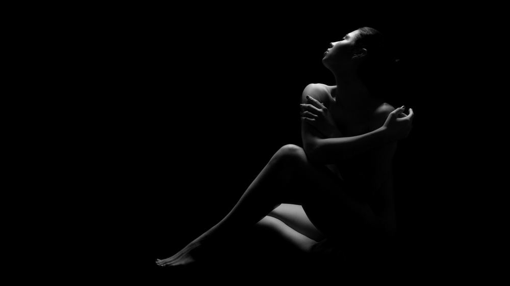 Nude woman sitting in darkness illuminated only by a single light source